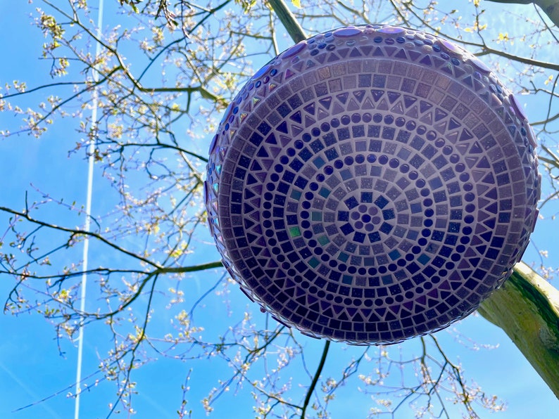 May include: A spherical garden ornament featuring a detailed mosaic pattern composed of small purple, blue, and dark violet tiles in circular, square, and triangular shapes, creating a geometric design across the entire surface of the object.