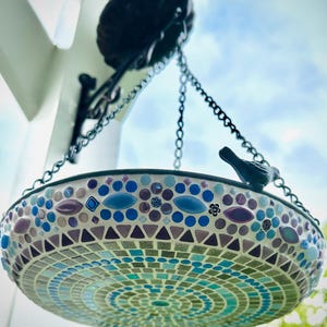May include: A hanging bird bath featuring a mosaic design with blue, purple, and turquoise tiles. The circular basin is suspended by metal chains from a black wall bracket and includes a small decorative black bird figure on the rim.