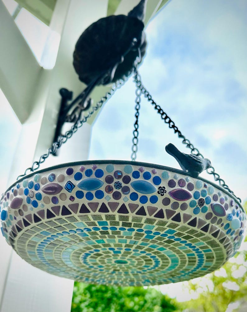 May include: A hanging bird bath featuring a mosaic design with blue, purple, and turquoise tiles. The circular basin is suspended by metal chains from a black wall bracket and includes a small decorative black bird figure on the rim.