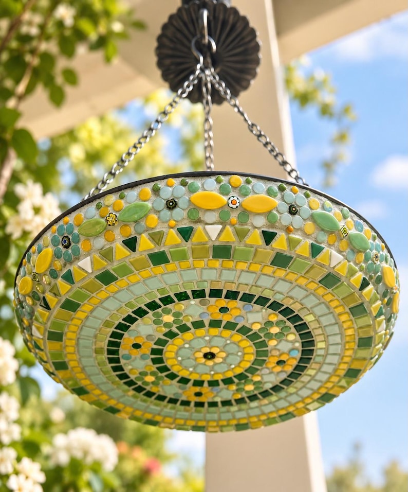 May include: A circular hanging bird bath featuring a mosaic design of green, yellow, and light blue glass tiles. The surface displays intricate floral patterns and geometric shapes, suspended by a dark metal chain attached to a central hook.