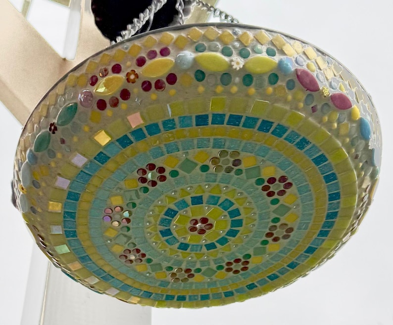 May include: A circular hanging suncatcher featuring a mosaic design composed of small glass tiles in shades of yellow, turquoise, light green, and red. The tiles are arranged in concentric circular patterns with floral motifs and a metal frame.