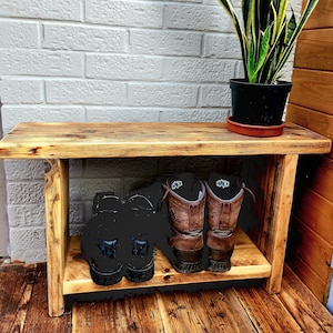 May include: A rustic wooden shoe bench with two shelves. The top shelf holds a potted snake plant. The bottom shelf displays two pairs of boots. The bench is made of light-colored wood and is placed against a white brick wall.