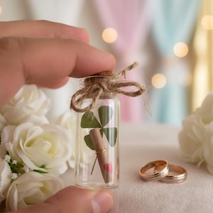 Puede incluir: Una pequeña botella de vidrio con un tapón de corcho atado con hilo, que contiene un trébol de cuatro hojas, un pergamino y un anillo. También se ven dos anillos de boda y rosas blancas, lo que sugiere un tema de boda o compromiso.