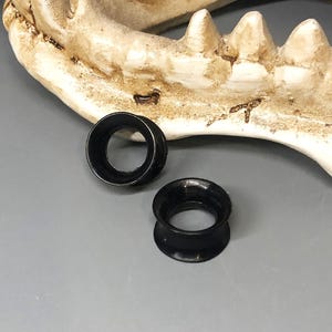 May include: Two black ear gauges are displayed against a gray background, with a portion of a bone structure in the background. The gauges are circular with a flared design, showcasing a sleek, minimalist aesthetic. The gauges are made of a shiny material.