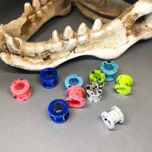 May include: Assortment of colorful acrylic ear gauges with black splatter paint designs. The gauges are in various sizes and colors, including pink, blue, green, and white.