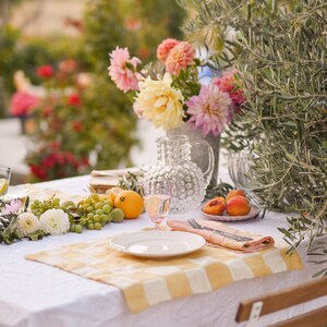 Puede incluir: Un mantel blanco con un mantel individual a cuadros amarillo y blanco. Un plato blanco, un vaso rosa y un tenedor de plata están colocados en el mantel individual. Una jarra de vidrio transparente con superficie texturizada está llena de agua. Un ramo de flores rosas y blancas está en un jarrón de metal detrás de la mesa. Uvas verdes y limones están sobre la mesa.