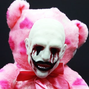 Op de afbeelding: Een roze pluche teddybeer met een wit gezicht en rood geverfde ogen en mond. De beer draagt een roze lint om zijn nek en heeft een hartvormige patch op zijn voet.
