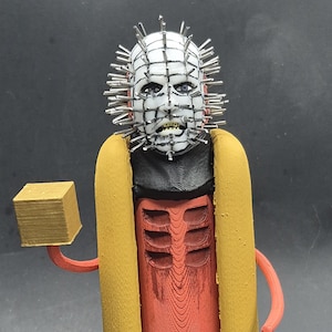 Puede incluir: Una escultura caprichosa impresa en 3D de un personaje de la película de terror Hellraiser, reinventado como un perrito caliente. La figura presenta una cara blanca con alfileres, una parte superior negra, un cuerpo de perrito caliente rojo, pan amarillo y sostiene un pequeño cubo dorado. Se encuentra sobre una base negra.