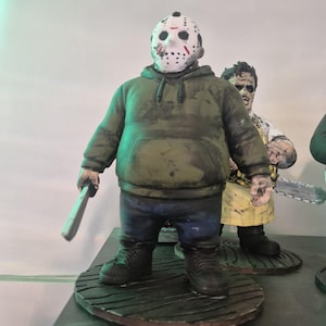 May include: A figurine of a character wearing a white hockey mask with red markings, a dark green hooded sweatshirt, and dark blue pants. The figure holds a silver-colored machete in one hand and stands on a circular base with a wood grain texture.