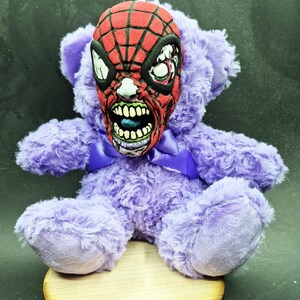 Puede incluir: Un oso de peluche morado con una máscara de Spider-Man roja y negra para la cabeza. La máscara tiene una cara de zombie verde y blanca debajo.