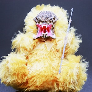 May include: A yellow plush toy with a predator-like face and a spear. The toy has a pink mouth and sharp teeth.