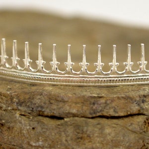 May include: A silver crown with a delicate, intricate design. The crown has a series of pointed arches with a decorative pattern.
