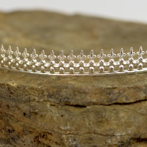 May include: A silver crown with a decorative fleur-de-lis design. The crown is made of metal and has a smooth, polished finish.