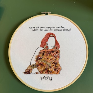 Könnte beinhalten: Gestickte Kunst in einem runden Holzreifen. Das Design zeigt eine stilisierte Figur mit roten Haaren und einem gemusterten Kleid. Der Text lautet: "let me ask you a very fair question, what do you do successfully? quickly."