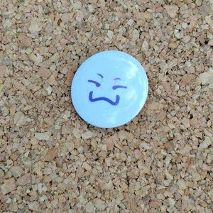 May include: A light blue button with a simple, smiling face drawn in dark blue ink. The button is set against a textured, brown corkboard background.