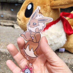 May include: A clear acrylic charm featuring a cartoon Eevee character in brown, cream, and pink. The charm is attached to a small bell with a pink bow. A plush Eevee toy is in the background.