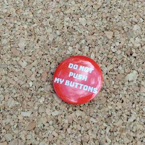 May include: A red button with the text "DO NOT PUSH MY BUTTONS" in white lettering. The button is round and sits on a textured, light brown surface.