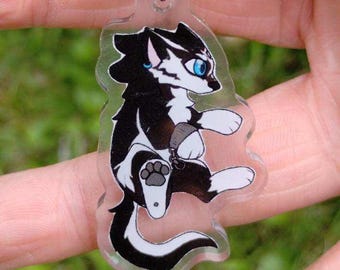 May include: A clear acrylic keychain featuring a cartoon skunk with black and white fur, blue eyes, and a gray belly. The keychain has a colorful iridescent clasp and a smaller charm with a cartoon character. The background is blurred green.