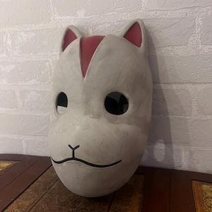 以下が含まれることがあります： 手作りのオフホワイトの猫のマスク。耳と額に赤いアクセントがあります。大きな目の穴、黒い鼻、笑顔の口が特徴です。コスプレやコスチュームイベントに適しています。