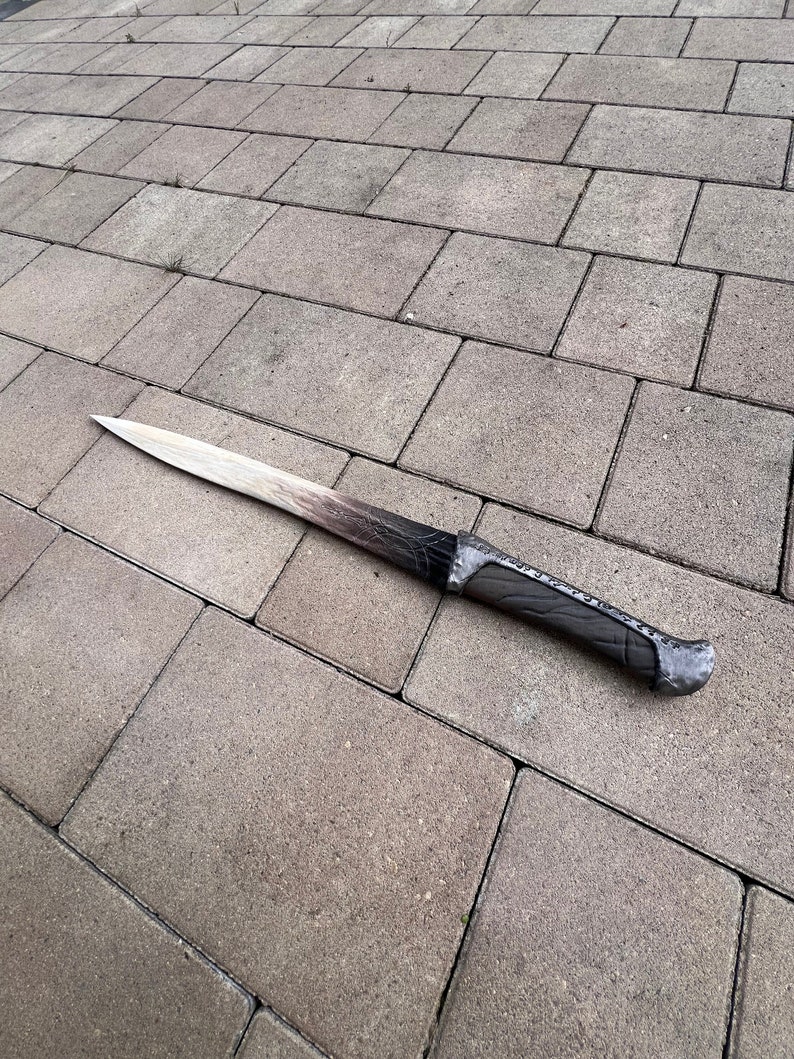 May include: A long, wooden sword with a black and grey handle. The sword is lying on a grey concrete surface.
