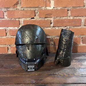 Könnte beinhalten: Ein schwarzer und silberner Helm mit einer verwitterten, abgenutzten Oberfläche. Der Helm hat ein detailliertes Design mit einer Reihe von Zähnen am unteren Rand. Zum Helm gehört ein passendes schwarzes und silbernes Unterarmstück.
