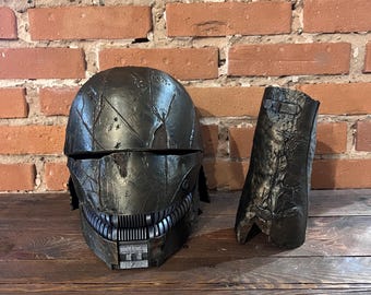 May include: A black and silver helmet with a weathered, distressed finish. The helmet has a detailed design with a series of teeth along the bottom edge. The helmet is accompanied by a matching black and silver forearm piece.