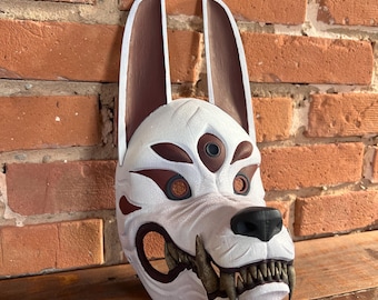 Op de afbeelding: Een wit en bruin hondenmasker met grote oren en scherpe tanden. Het masker heeft bruine accenten rond de ogen en op de wangen. De neus is zwart. Het masker wordt getoond tegen een bakstenen muur en een houten oppervlak.