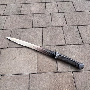 May include: A long, silver-colored sword with a black handle. The sword is lying on a gray concrete surface.