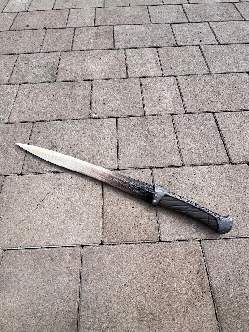 May include: A long, silver-colored sword with a black handle. The sword is lying on a gray concrete surface.