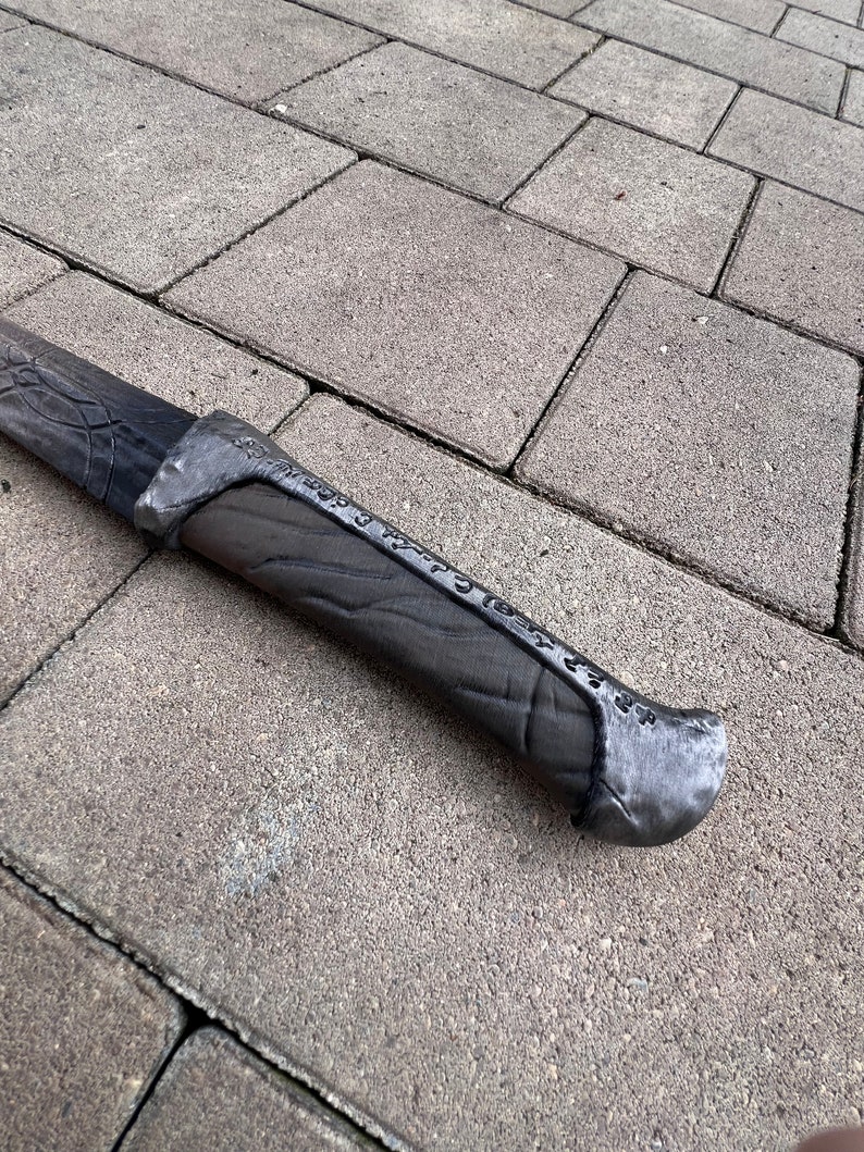 May include: A close-up of the handle of a silver-colored sword with a black, textured grip. The handle has a raised inscription that reads "One Ring to Rule Them All".