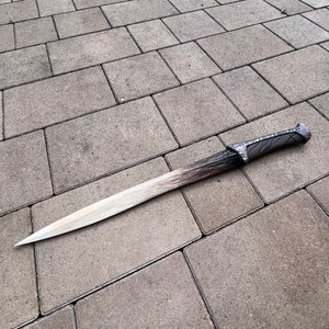 May include: A long, silver-colored sword with a black handle lies on a gray concrete surface.