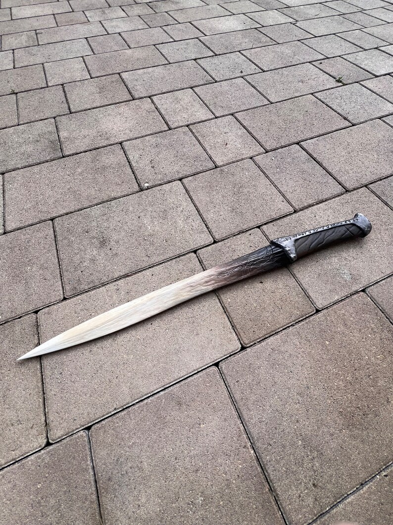 May include: A long, silver-colored sword with a black handle lies on a gray concrete surface.