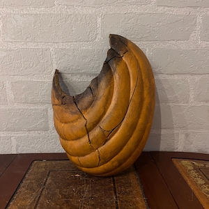 以下が含まれることがあります： 半月型の彫刻装飾品。暖かく金褐色の色合いで、より暗く焦げた部分があります。表面は質感のある畝のある模様で、木材で作られているようです。