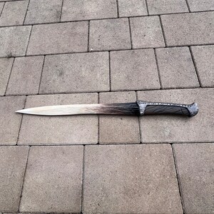 May include: A long, silver-colored sword with a black handle and intricate details. The sword is lying on a gray concrete surface.