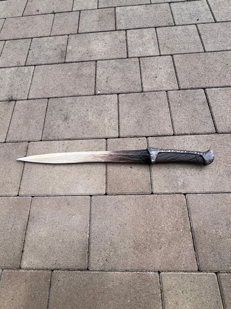 May include: A long, silver-colored sword with a black handle and intricate details. The sword is lying on a gray concrete surface.