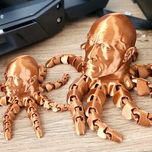 May include: Two 3D printed bronze-colored octopus sculptures with human heads. The larger octopus has eight tentacles and the smaller octopus has six tentacles.