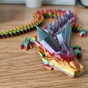 May include: A rainbow-colored 3D printed dragon figurine with a detailed, faceted design. The dragon is posed with its head raised and its tail curled.