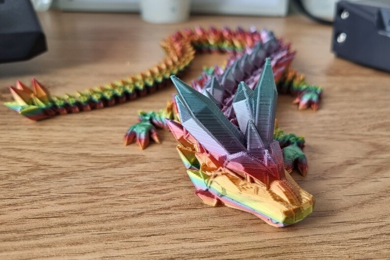 May include: A rainbow-colored 3D printed dragon figurine with a detailed, faceted design. The dragon is posed with its head raised and its tail curled.