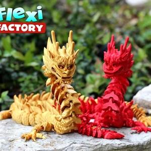 May include: Two articulated dragon figurines, one gold and one red, posed on a rock. The dragons are made of plastic and have a segmented body. The text "Flexi Factory" is visible in the background.