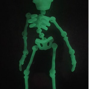 May include: A glow-in-the-dark skeleton figurine. The skeleton is posed standing with its arms outstretched. The figurine is made of a green plastic material.