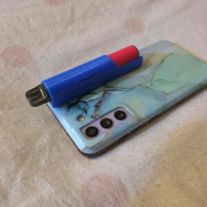 K&ouml;nnte beinhalten: Ein blau-roter Vape-Pen liegt auf einem Smartphone mit einer hellblau-gr&uuml;nen Marmordesign-H&uuml;lle. Das Telefon hat drei Kameralinsen und einen Blitz. Der Hintergrund ist ein wei&szlig;er Stoff mit rosa Punkten.