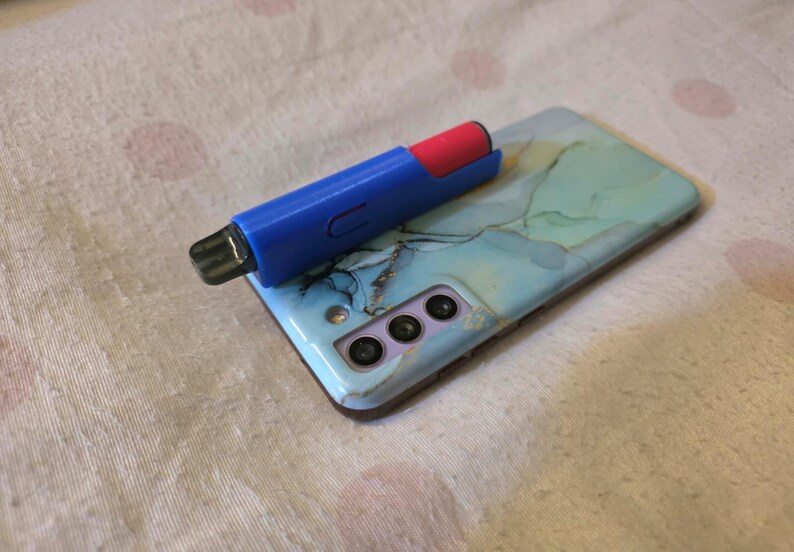 K&ouml;nnte beinhalten: Ein blau-roter Vape-Pen liegt auf einem Smartphone mit einer hellblau-gr&uuml;nen Marmordesign-H&uuml;lle. Das Telefon hat drei Kameralinsen und einen Blitz. Der Hintergrund ist ein wei&szlig;er Stoff mit rosa Punkten.