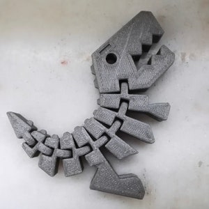 Puede incluir: Un juguete de esqueleto de dinosaurio de color gris impreso en 3D. El dinosaurio est&aacute; en posici&oacute;n de pie con la boca abierta.