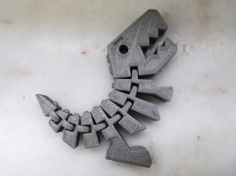 Puede incluir: Un juguete de esqueleto de dinosaurio de color gris impreso en 3D. El dinosaurio est&aacute; en posici&oacute;n de pie con la boca abierta.