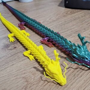 May include: Two colorful dragon figurines, one yellow and one with iridescent green and purple scales. The dragons have long, flowing tails and are posed with their heads raised.