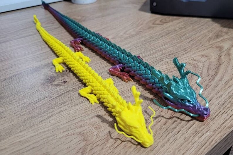 May include: Two colorful dragon figurines, one yellow and one with iridescent green and purple scales. The dragons have long, flowing tails and are posed with their heads raised.