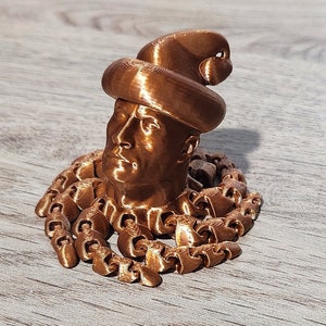 Puede incluir: Una escultura de cabeza humana con sombrero, impresa en 3D en color cobre. La cabeza est&aacute; rodeada por una cadena.