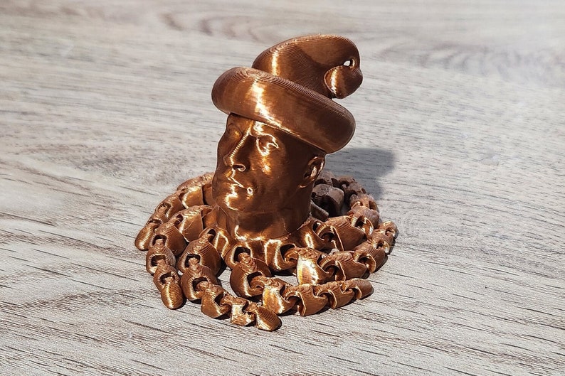 Puede incluir: Una escultura de cabeza humana con sombrero, impresa en 3D en color cobre. La cabeza est&aacute; rodeada por una cadena.