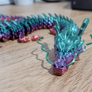 May include: A 3D printed dragon figurine with a gradient of pink, purple, teal, and green. The dragon has a long, serpentine body and a detailed head with a curled whisker.
