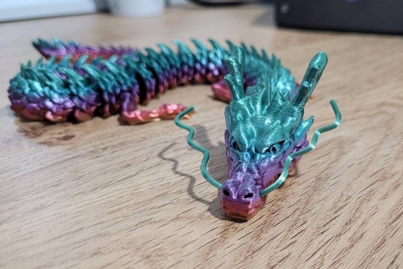May include: A 3D printed dragon figurine with a gradient of pink, purple, teal, and green. The dragon has a long, serpentine body and a detailed head with a curled whisker.